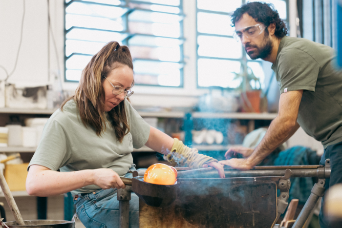 Artists Fiona Byrne and Aria Kiani working in the hot glass studio. Fiona is shaping molten glass with a wooden tool. 