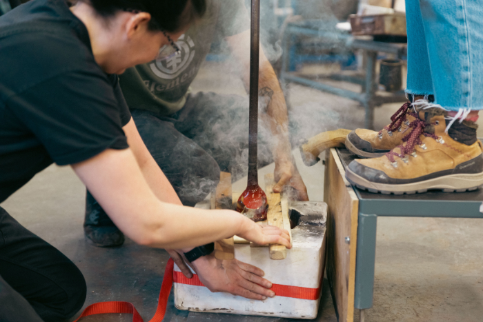 Artist Fiona Byrne stands on a step blowing down a glassblowing pipe pushing molten glass into a plaster mould held by Aria Kiani and Laura Quinn.