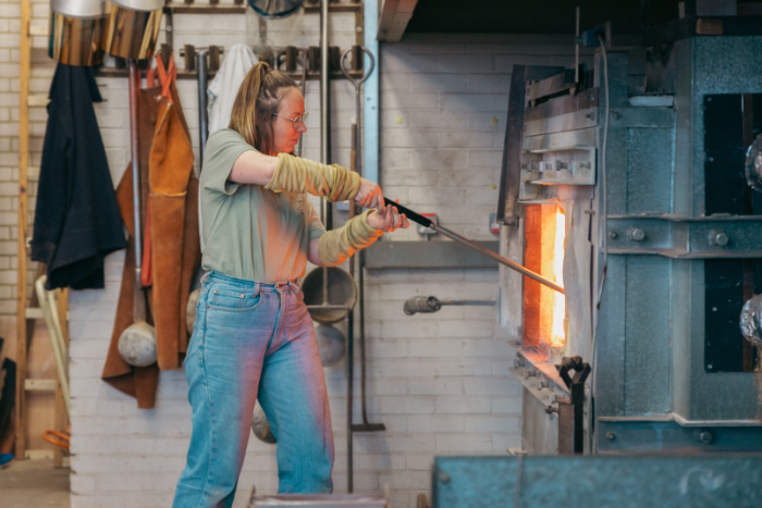 Artist Fiona Byrne works at a glass furnace. She has a long metal pipe which she is dipping in to the molten glass.