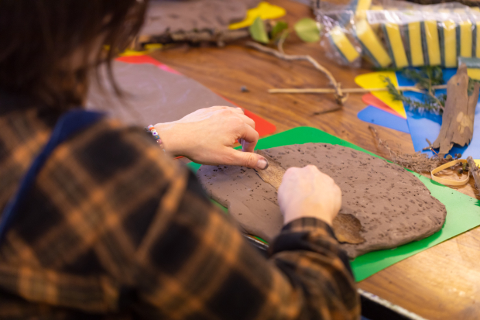 Workshop participant pressing a handmade tool into a roughly square clay tile. The clay is a deep red brown colour.