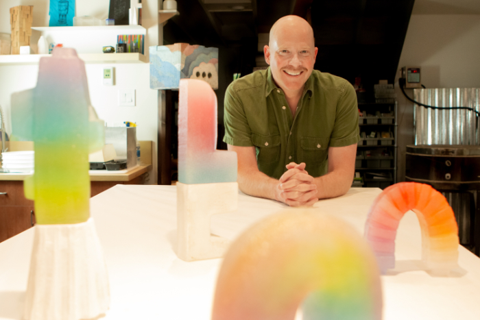 Glass artist Morgan Madison seated at a table behind several of his kiln-cast rainbow-coloured abstract glass forms.
