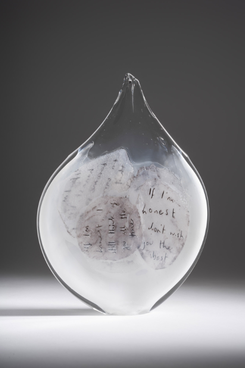 A clear and white glass vessel with a narrow top containing handwritten messages on pate de verre glass.