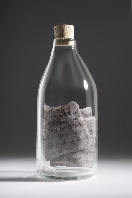 A clear glass bottle containing squares of pate de verre glass with handwritten notes on them.