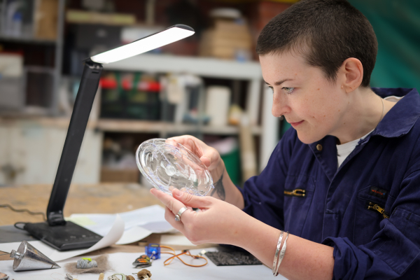 Glass artist Sophie Longwill sitting in the studio at the workbench examining a piece of round, clear glass .