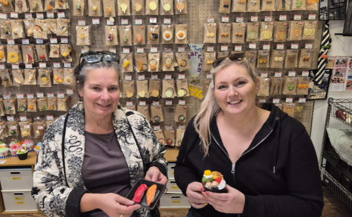 Glass artists Karen Browning and Sarah Brown hold their fake food creations that they made in Osaka, Japan. Behind them is a wall of pieces of fake food in display packaging.