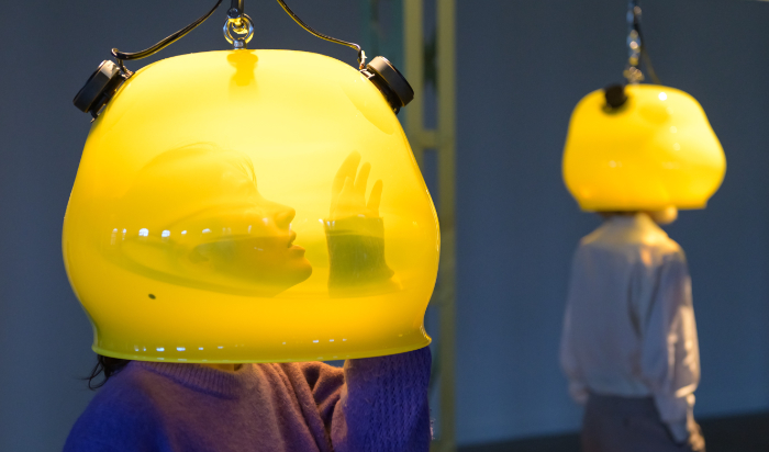 Inverted large yellow glass pods suspended from above which a person can put their head inside to hear recorded sounds. Installation made by Stef Veldhuis.