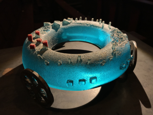 This donut-shaped sand-cast glass piece has small shapes on the top and three steel wheels. 