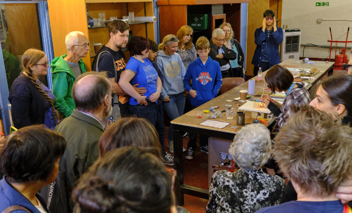 Glass artist Yukiko Sugano seated at a long table demonstrating her flame working technique to a crowd of onlookers at the International Festival of Glass 2024 in the UK.