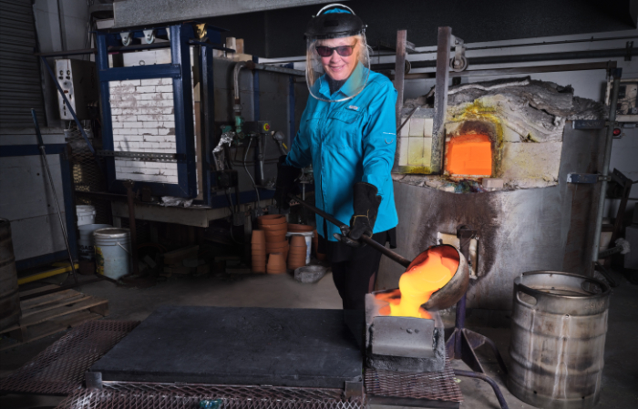Trish Duggan wearing a fireman's mask and holding a large ladle to pour molten glass into a mould at Sigma Studio.