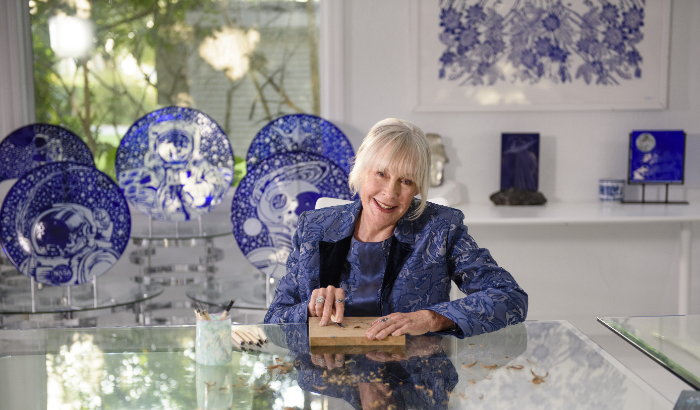 Trish Duggan carving woodblock seated at a table with assorted blue-and-white glass panels on display in the background.