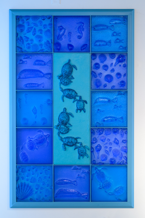 A vertically rectangular blue glass panel made up of lots of squares of glass with different underwater themes. In the centre is a smaller vertical rectangle panel featuring swimming turtles.