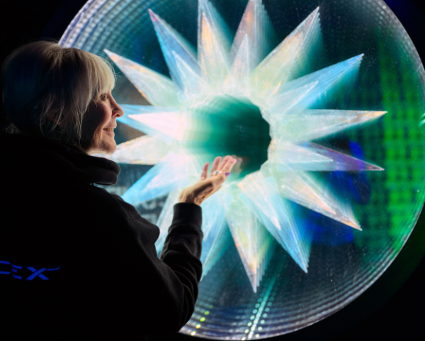 Trish Duggan rear view pointing out a large layered glass artwork in front of her called 'Star Gate Exit Portal Planet Earth' which looks like a white star shape with a hole in the centre suggesting a tunnel.