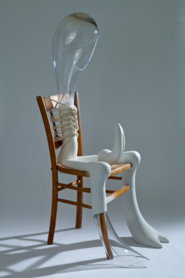 Mixed media figure tied on a wooden chair with clear glass head shape and white leg-shapes.