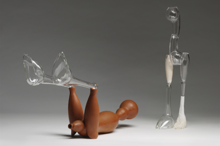 Mixed media sculptures of two figures using glass. One stands with no arms and the other lays down on its back with legs raised in a giving birth position.