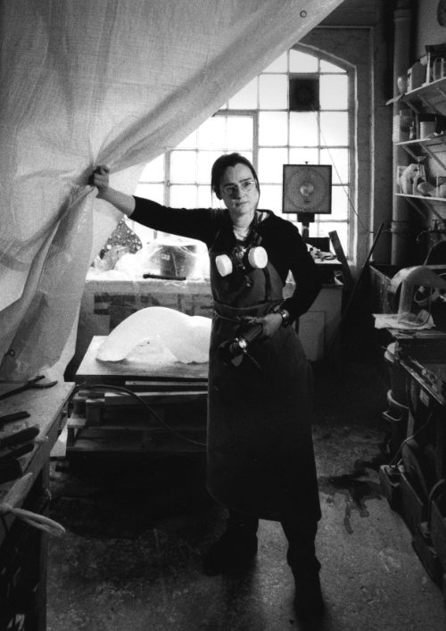 Emma Woffenden black and white image in studio where she stands holding back a large curtain with a big window in the background and a table made of pallets and shelves of work on display.