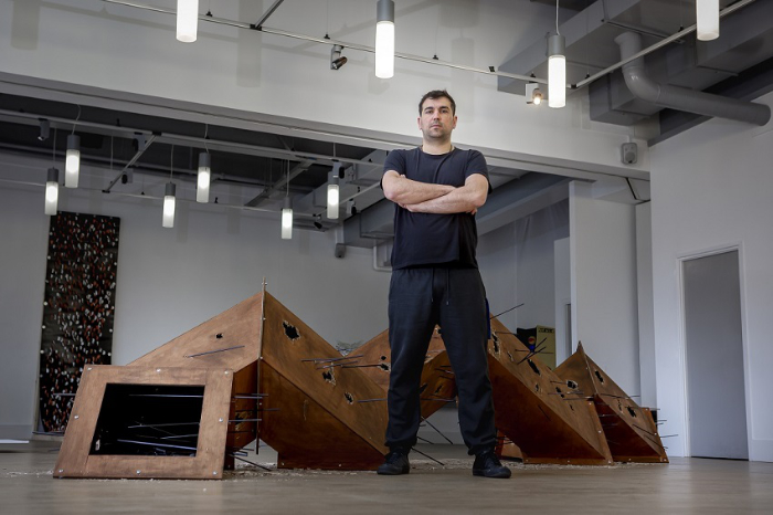 Antonis Koutouzis at his degree show, University of Sunderland, standing in front of his large sculpture of squared and angled brown blocks.