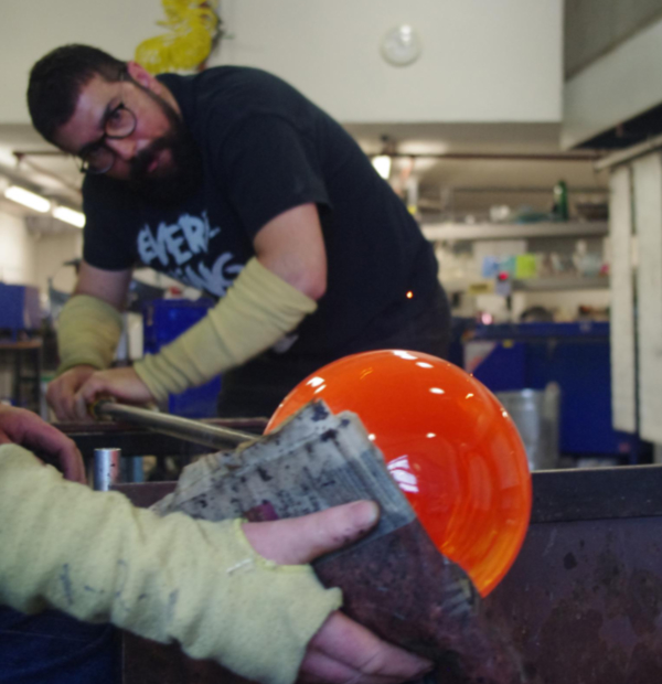 Antonis Koutouzis working on the glassblowing process. He turns a blob of molten glass on a long pole while a hand in the foreground shapes it with newspaper. 
