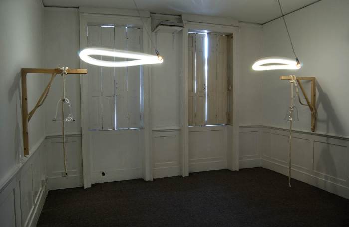 Installation project in room with two swinging lightbulbs in circular motion and suspended bell-shaped glass pieces suspended from wooden brackets at each side all in a room with white walls.