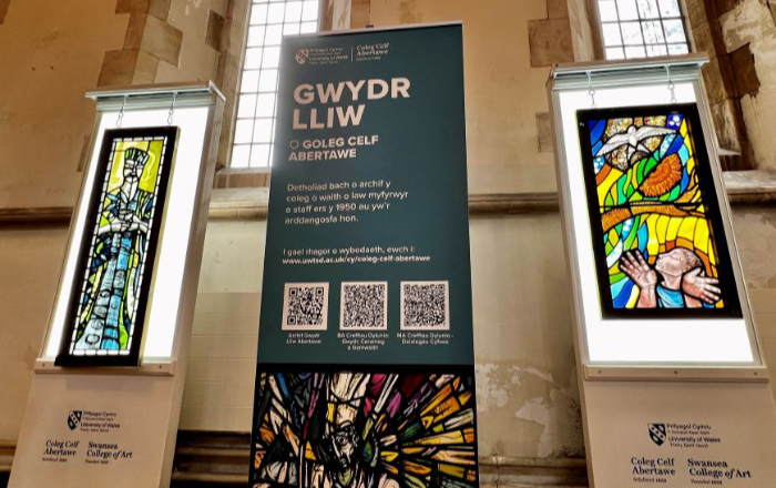 Two stained glass panels hanging in front of illuminated light boxes in a church setting with a Welsh language information panel standing between them