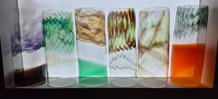 Six mouth blown glass cylinders standing vertically in a row against a lit background to show off the bold solid and delicate patterns and colours.