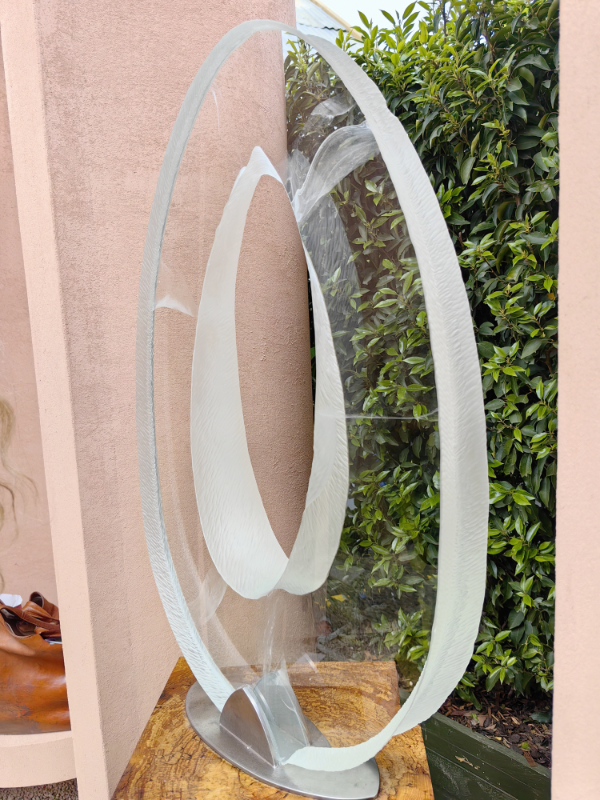 A large garden sculpture made from clear glass in the shape of an elongated O, frosted around the outside edge, which is an uneven depth.
