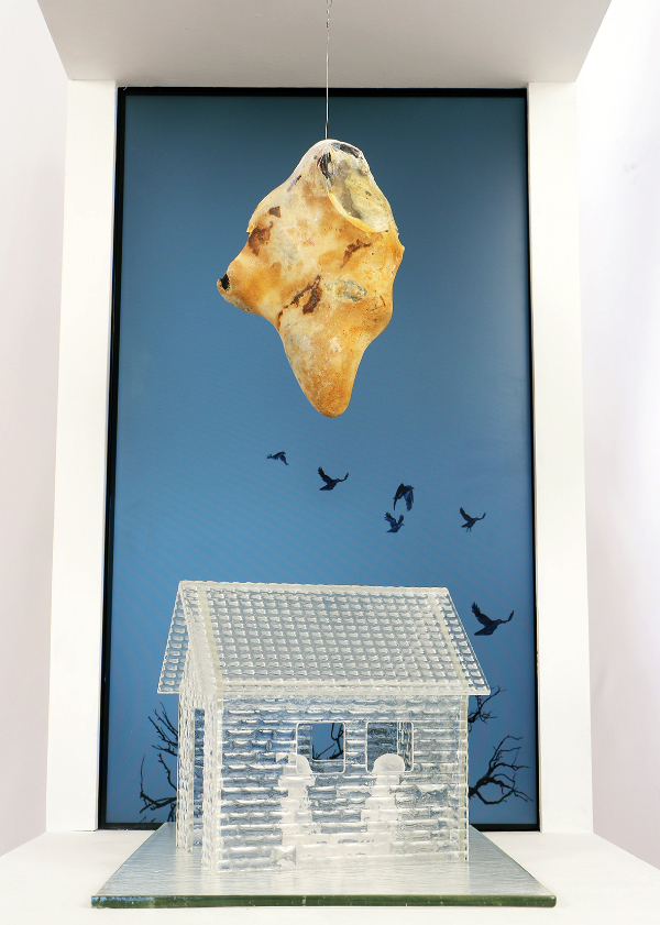 Installation featuring a screen of flying birds on blue in the background with a 'Sword of Damecles' pointed rock hanging above a clear glass house with figures inside. Made from Waterjet cut, fused, and cast glass, animation, flint rock, Raspberry Pi microcomputer.