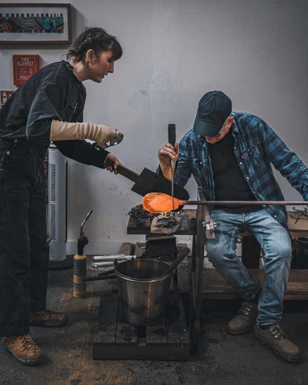 Tuesday assistant Saskia Dammers stands with paddle shaping molten glass for Mark Fenn who is seated and shaping the molten glass with large metal implement while it is attached to the end of a long metal blowing pipe.