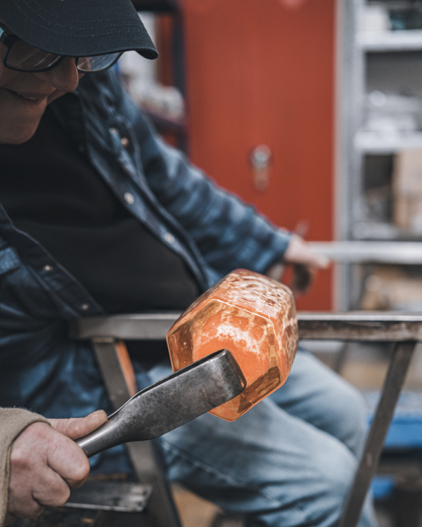 Mark Fenn using a metal tool to shape molten glass on the blow pipe that has come from a hexagonal mould.