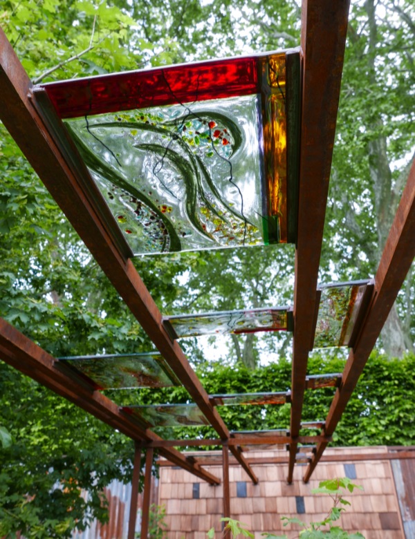 Close-up of a fused glass panel mounted on mouthblown glass held in a rusted metal beam structure above the planting. 