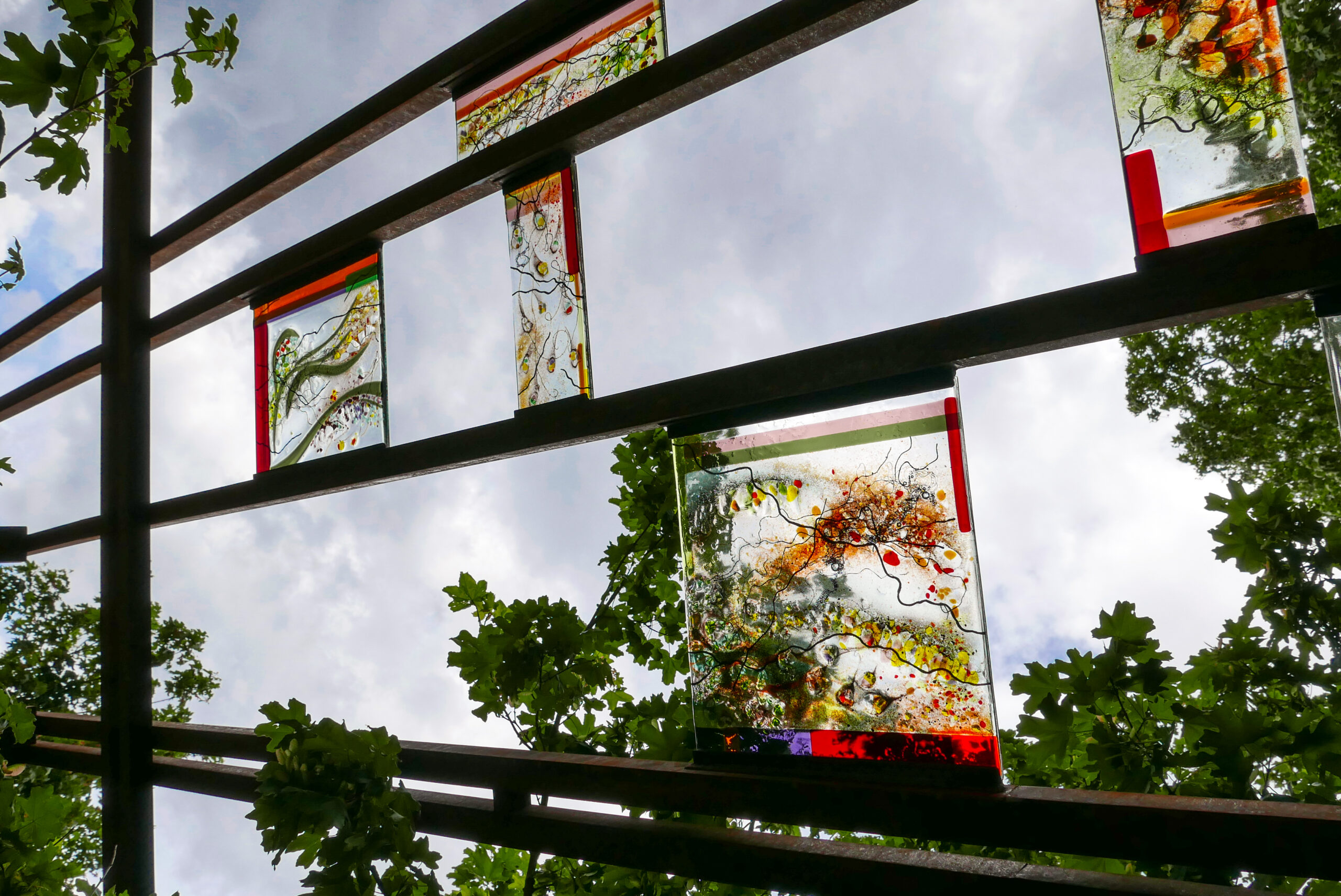 View of rectangular decorative fused and mouthblown glass panels mounted in metal beams with sky and tree branches in the background.