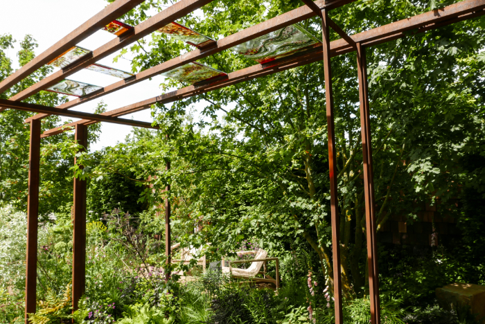 A view across the garden showing horizontal metal beams supporting decorative glass panels and seating surrounded by soft planting. 