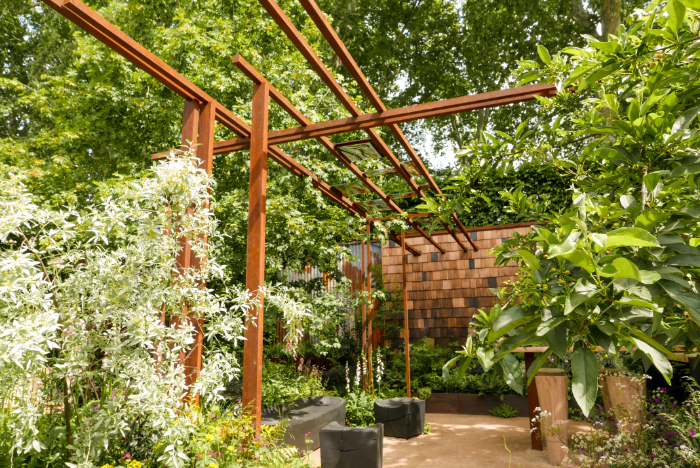 A view across the show garden at Chelsea featuring recycled materials including metal bars and tiled wall among soft planting. Decorative glass rectangular panels are fitted between the beams. 