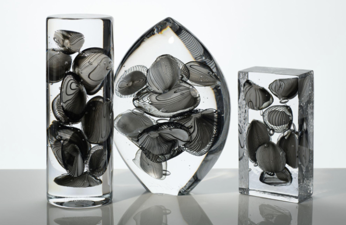 Three glass sculptures in different shapes with black and grey inclusions within clear polished glass. All made by Peter Layton