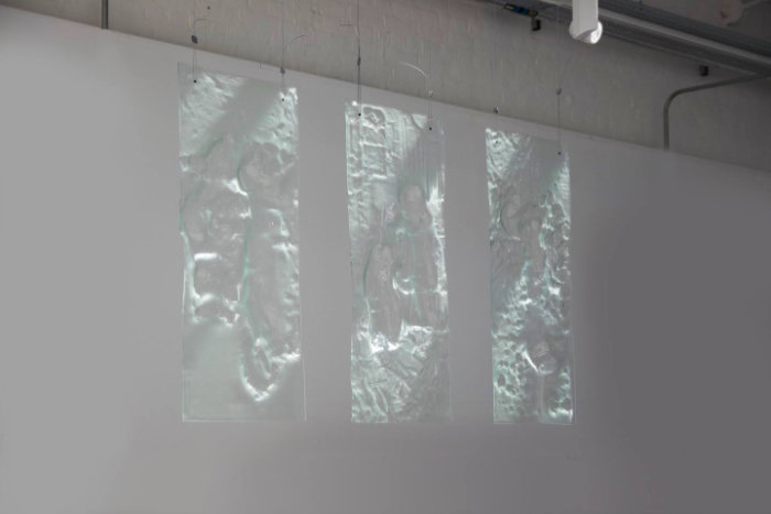 Three vertical glass artworks in slumped float glass hang in an exhibition space against a white wall.