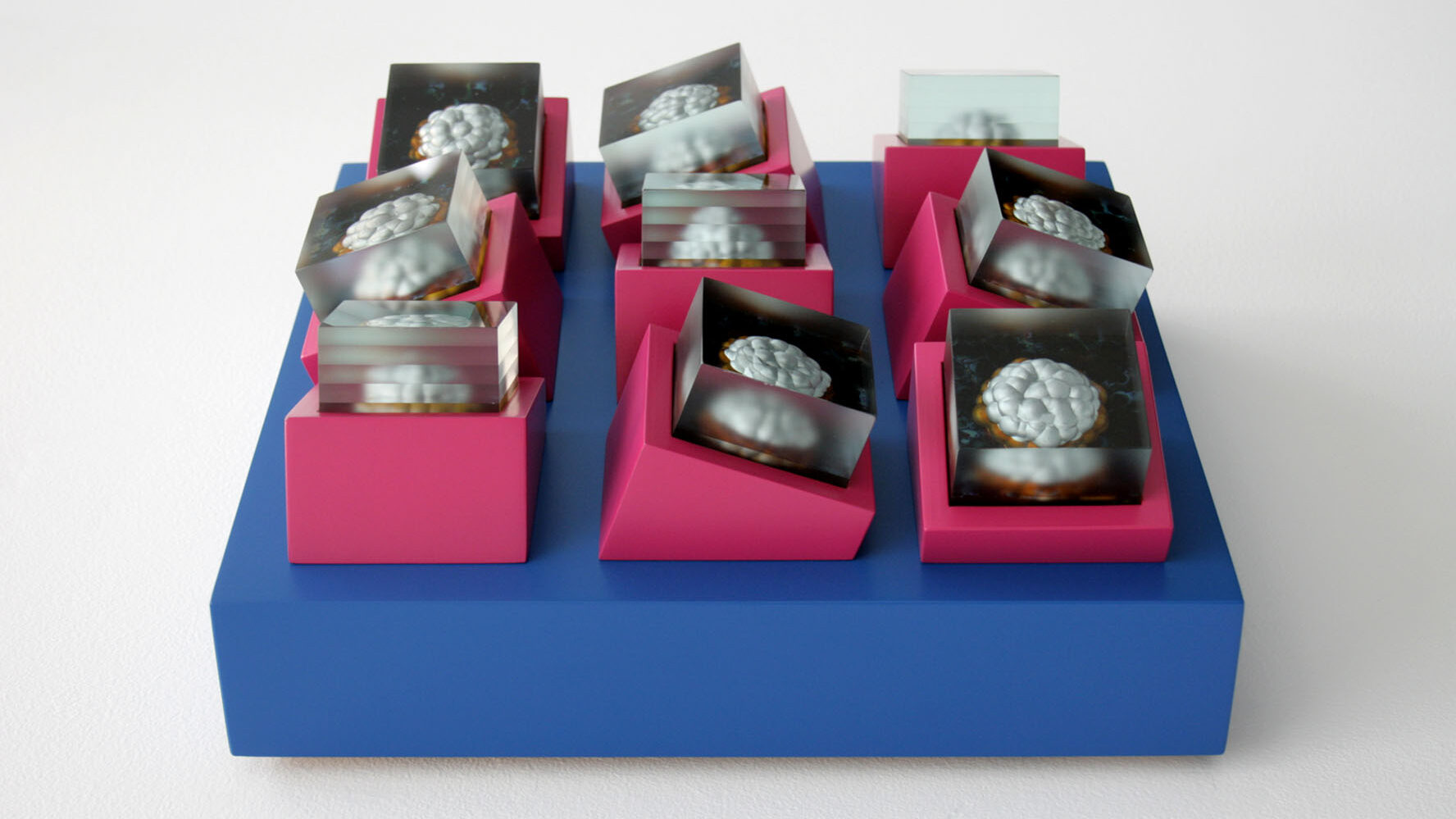 A mid blue square sided shallow box supporting small cubes of cut and polished glass layers in angled pink cubes. All made by Stacey Poultney and this sculpture is titled Urban Echoes.