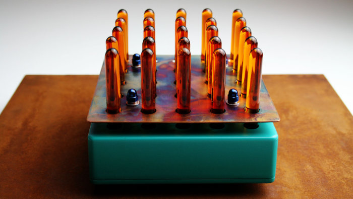 A square-based box in green with a layer of metal on top supporting 5 x 5 amber glass tube shapes pointing vertically in this industrial sculpture.