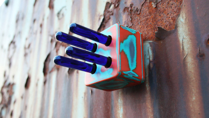 Wall sculpture of cube shape in orange and blue with four dark blue tube shapes sticking out. It is mounted on a rusty metal fence.