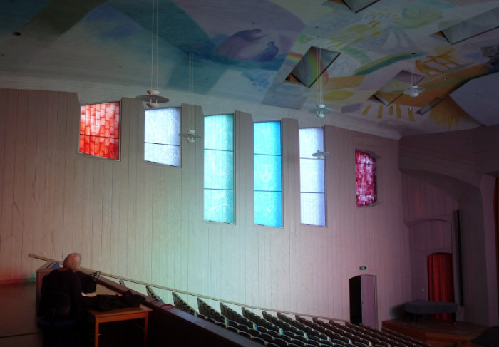 A view across chairs in Sweden's Ytterjarna auditorium to the north side wall featuring three short and three long carved glass window panels, each in a different monotone glass.