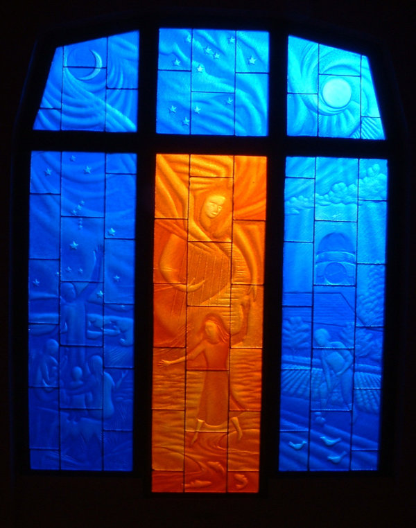 'The incarnation child' window features three long vertical carved stained glass panels with three smaller ones above. Five are in blue monochrome and the centre bottom one is in vibrant orange. It resides at the Michael Hall Steiner School in Forest Row, East Sussex.