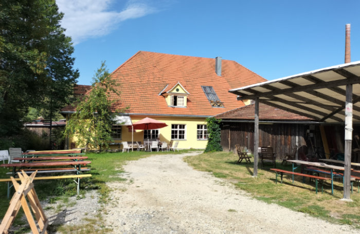A view along a gravel track towards a yellow rustic building in Germany with a terracotta coloured roof. Picnic benches are either side of the track and the right ones have a weatherproof cover built over them.