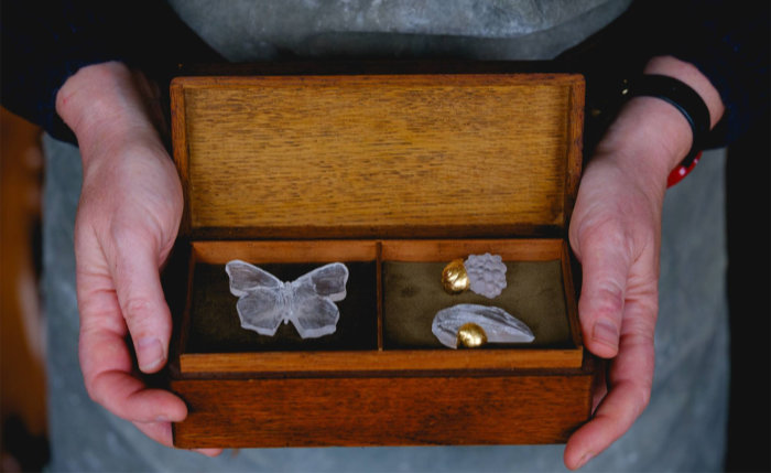 Close-up from above of hands holding either side of a wooden box holding a clear glass cast butterfly, fruit and fish with gold embellishments. Item by Effie Burns and titled "land sea sky".