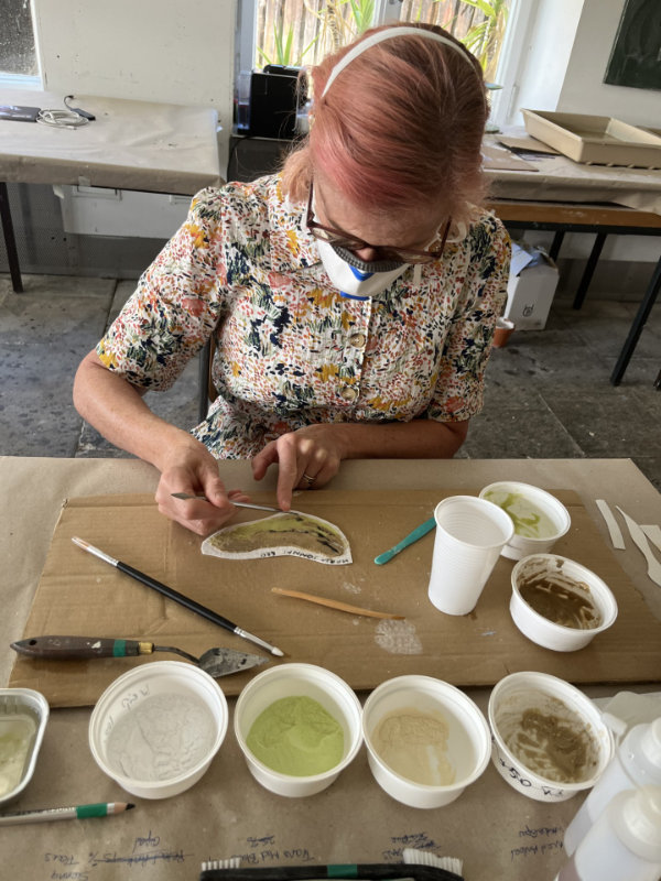 Maria Donai seated at a workbench facing work on the table wearing a facemark and with pots of glass frit colours in the foreground.