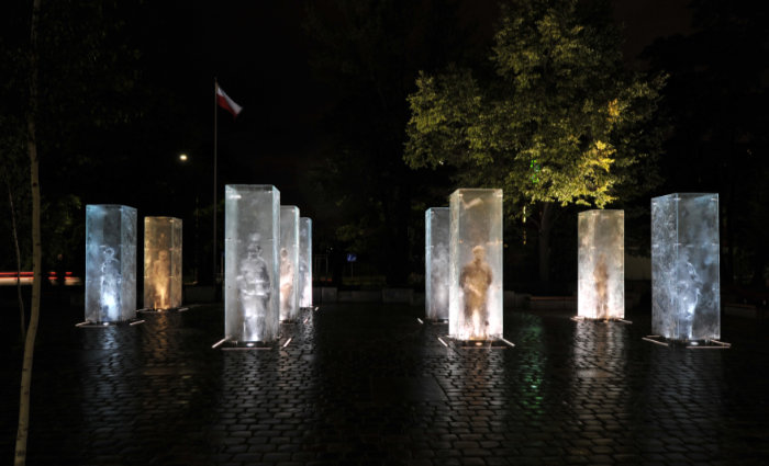 9 cuboid clear cast glass vertical structures with cast glass life-sized figures inside representing anti-Soviet partisans. It is a glass installation in a city square in Poland and this image is taken at night so the cuboids are lit inside and with strip lights around each one. They stand in a military square formation.