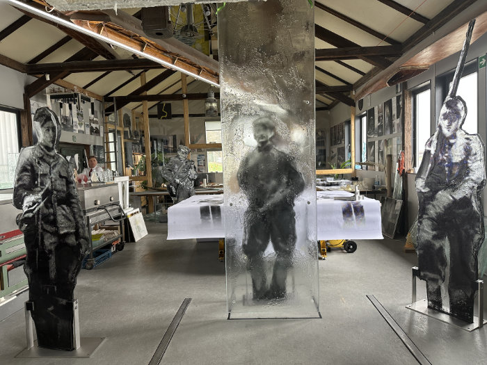 A view across a spacious art studio showing two cast glass partisans held upright by metal stands and another encased in a clear, textured glass cuboid.