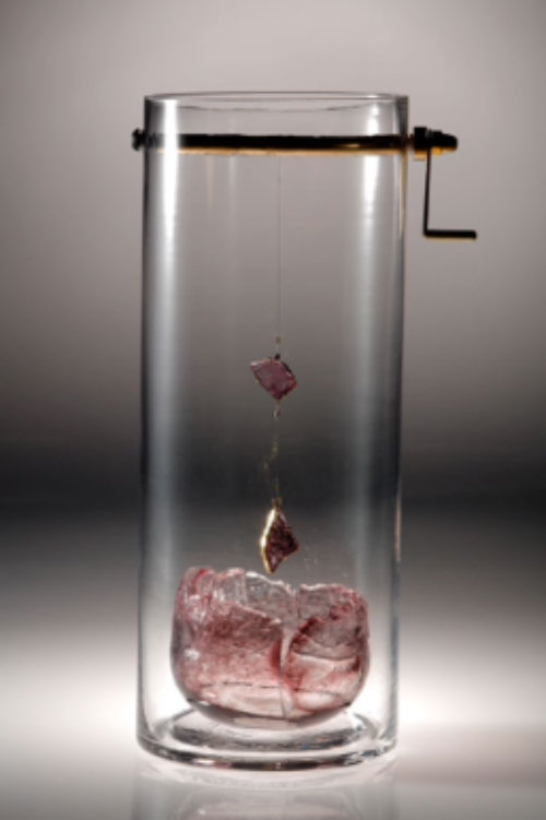 Vertical clear glass cylinder with a metal rod inserted through it near the top and a small handle on one side. Two angular shaped pale pink glass pieces are suspended on thin wire from the rod above an organic dish shaped piece of pale pink glass sitting in the base of the cylinder. By Cynnie Leung.