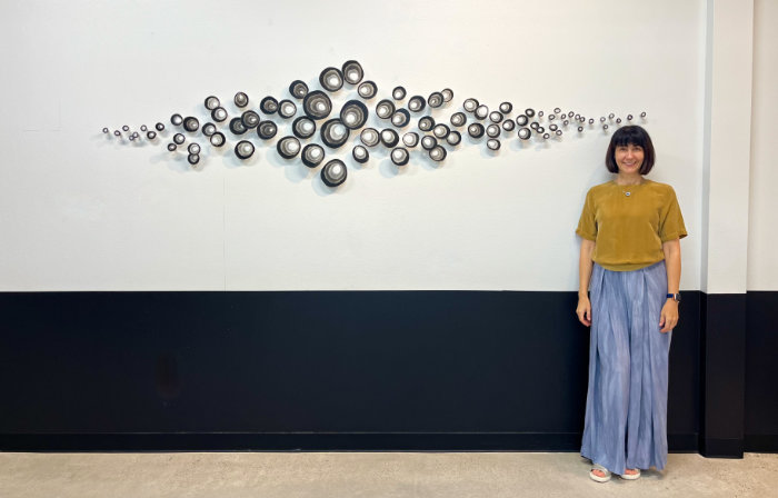 Glass artist Malina Cipleu stands to the right of an installation of cast glass organics rounded shapes in different sizes in tones of grey mounted on a wall and spanning a width of about 2 metres. The main collection of larger pieces is concentrated in the centre, with gradually smaller pieces spreading out to left and right in a tapering formation.