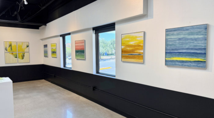 A view across an art gallery with 6 kiln-formed glass square artworks mounted on the walls and two windows between them.