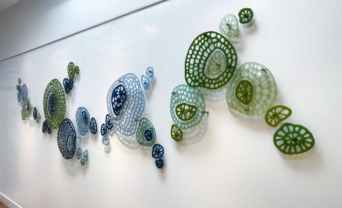 Wide wall art installation of tones of blue and green fused glass organic, rounded shapes with holes in called Primeval Gathering.