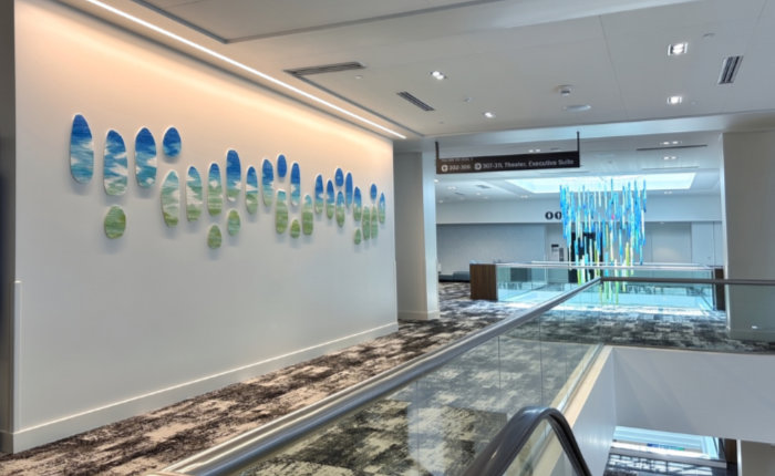 A view across to a white interior wall in Dayton Convention Center in Ohio, USA, featuring a run of kiln-cast glass ovals positioned organically horizontally along the walls. They are blue and white at the tops and green at the bottoms, suggesting sky and land. In the foreground is a patterned brown carpet and top of an escalator and through an opening at the rear of the image there is an aqua blue vertical hanging sculpture whose details cannot be made out.