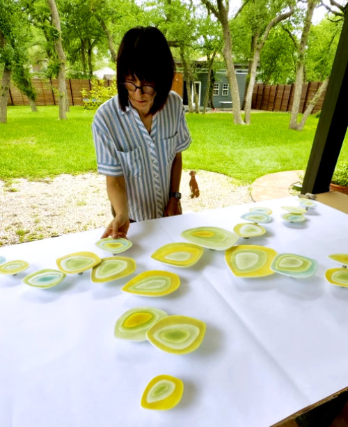 Artist Malina Cipleu stands at a large table with a white cover with a garden in the background. On the table she arranges about 20 yellow toned organic oval shaped cast glass pieces into a spreading pattern.